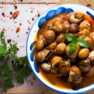 Plato de caracoles en salsa tradicional servidos en cuenco de cerámica, acompañados de hierbas aromáticas y especias, representación de una receta clásica de la gastronomía española.